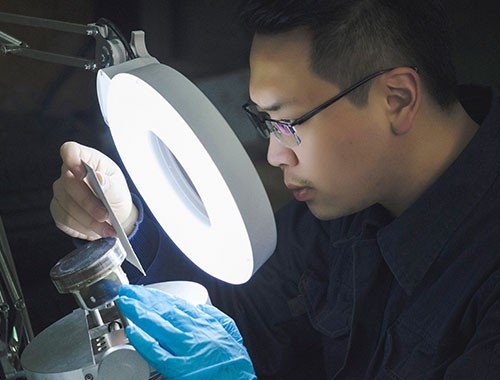 Worker using ring light and gauge tool to rate an automotive engine part in a lab.